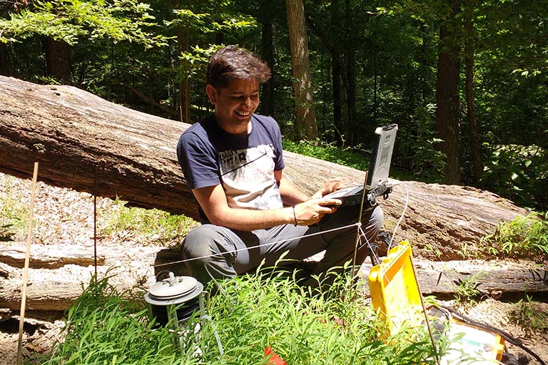 A man leans against a fallen log as he enters data from the research plot into his laptop.