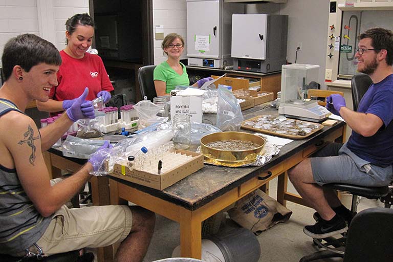 Four lab members work around a table in the lab cluttered with plastic bags filled with soil, test tubes, and instruments.