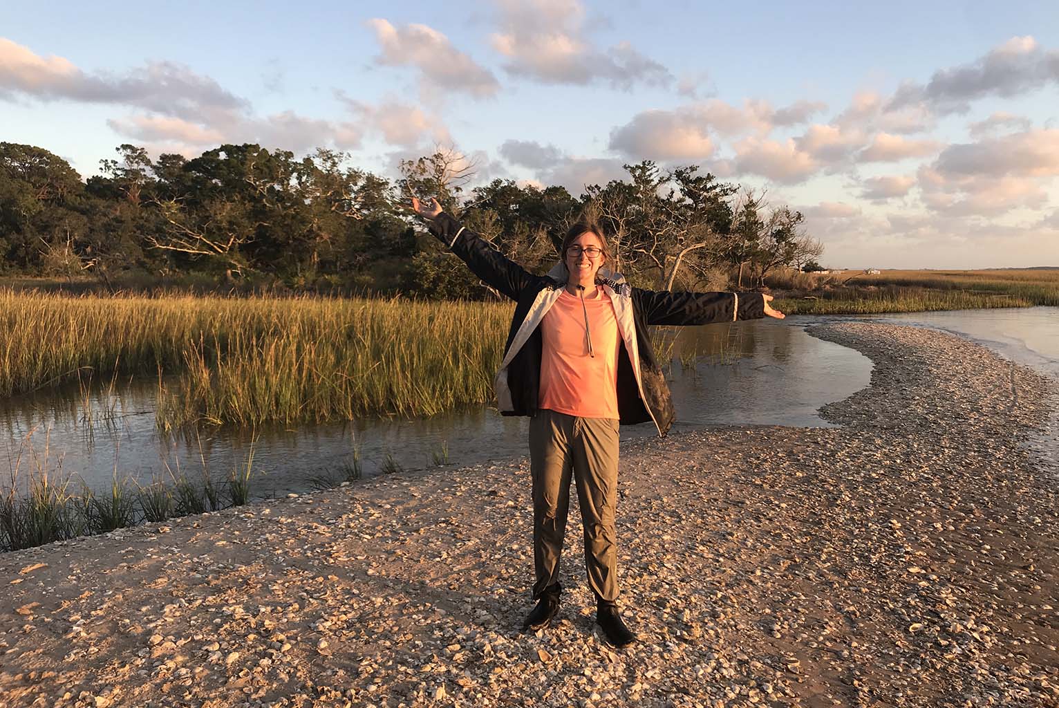 Nicole Spanier wears a light-weight jacket while standing on a dry strip of gravel-covered soil with arms outstretched. Flooded grassland surrounds her. Green-leafed trees are in the background. The sky is pale blue with clouds tinged in pinks and grays, suggesting a sunset.